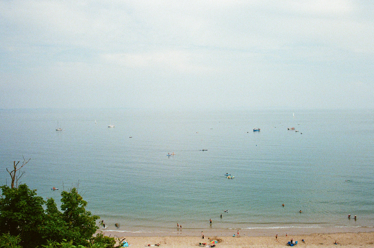 A scenic beach and ocean view with calm blue-green water, a sandy shoreline, small boats on the sea, and a few people near the beach. Trees are visible in the lower left corner under a hazy sky.