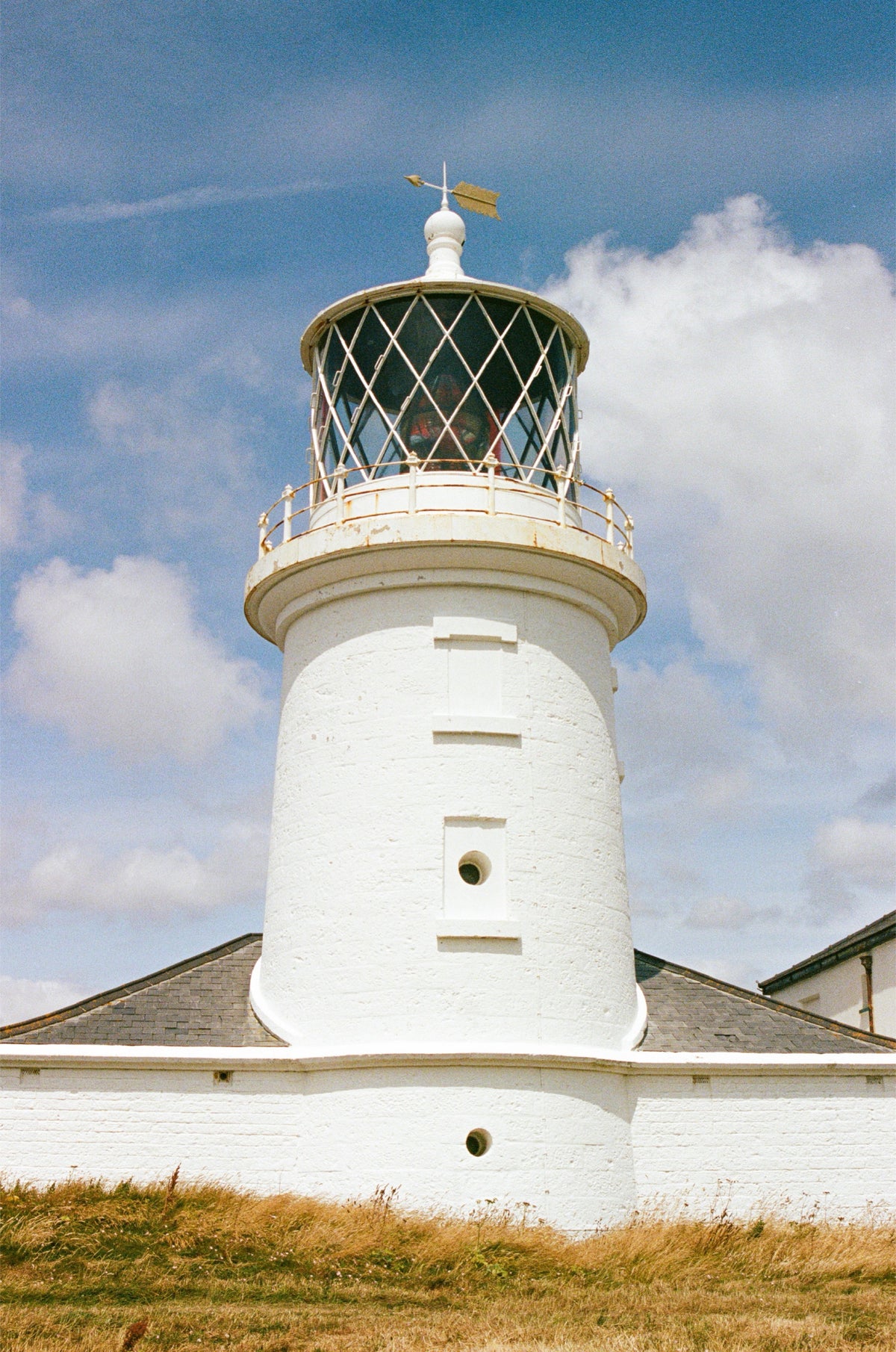 A white lighthouse with a glass lantern room and small keeper's cottage, photographed outdoors against a blue sky with clouds and dry grass in the foreground.