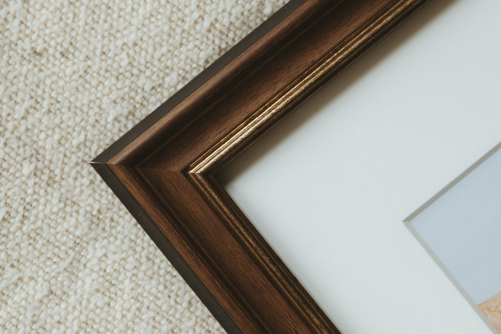 Close-up of a dark brown wooden picture frame corner with a white mount, placed on a textured beige carpet.