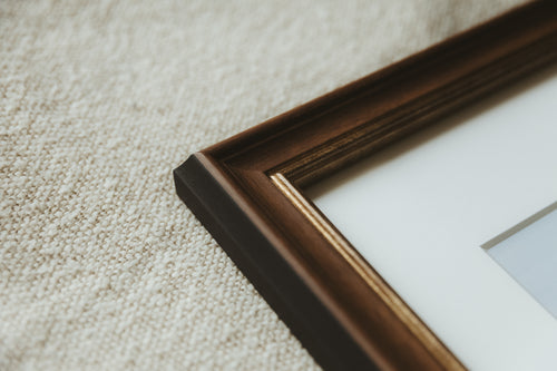 Close-up of a dark brown wooden picture frame corner with a white mount, resting on a textured beige fabric background.