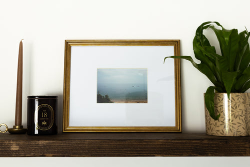 A framed landscape photograph with a white mount and a gold frame, styled on a dark wooden shelf next to a brown taper candle, a black candle tin, and a potted green plant against a white wall.