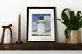 A framed art print of a white lighthouse under a blue sky, styled on a dark wooden shelf next to a potted green plant, a brown candle, and a white decorative jar with a brown ribbon.