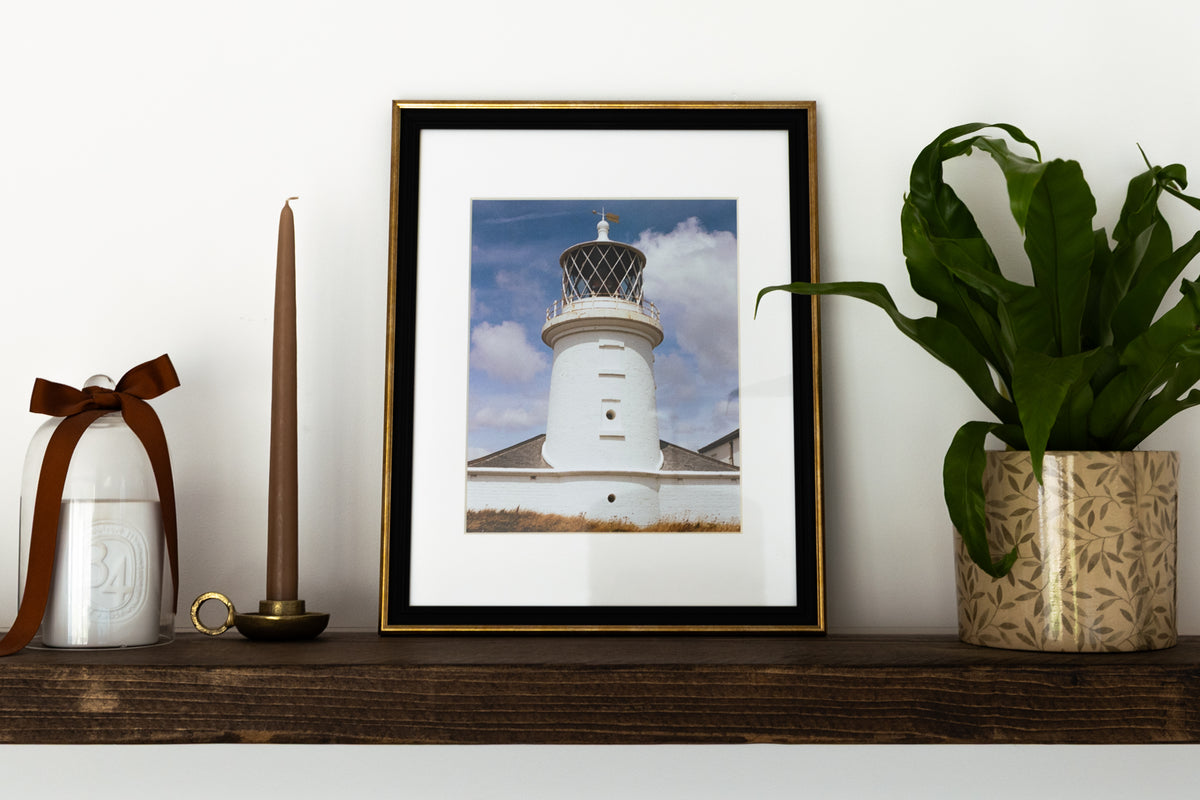 A framed art print of a white lighthouse under a blue sky, styled on a dark wooden shelf next to a potted green plant, a brown candle, and a white decorative jar with a brown ribbon.