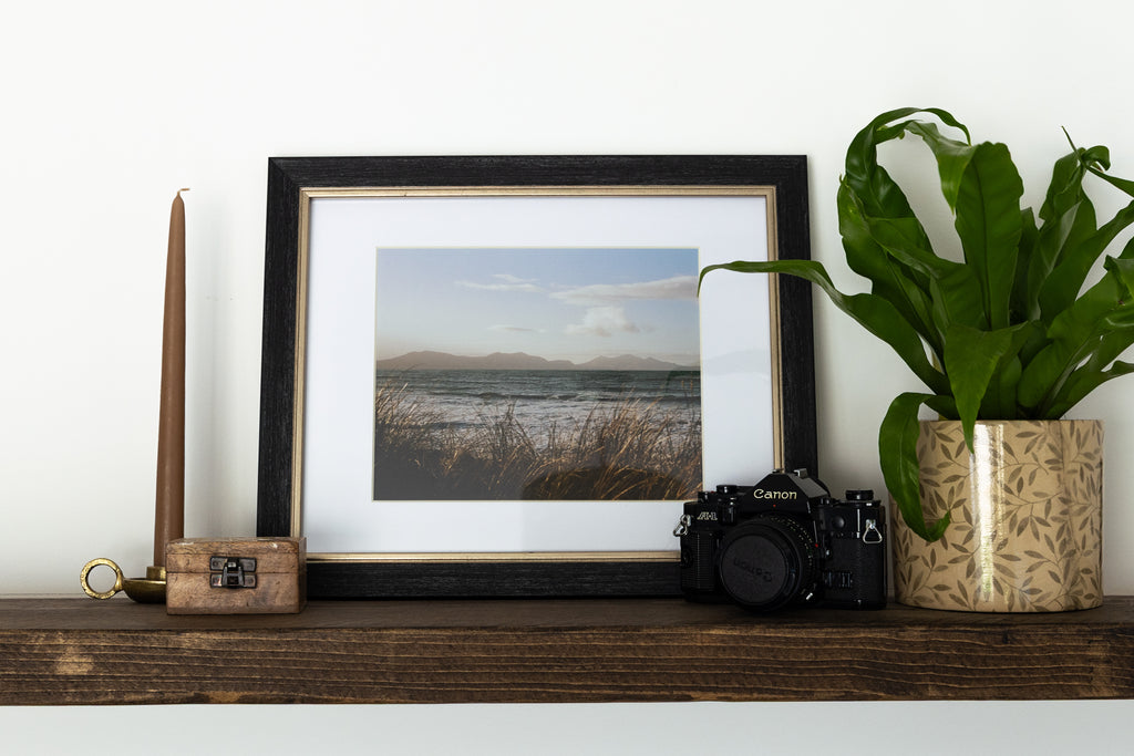 A framed landscape photograph of a beach and distant mountains, styled on a wooden shelf next to a black camera, a tall wooden candle holder, and a potted green plant against a white wall.