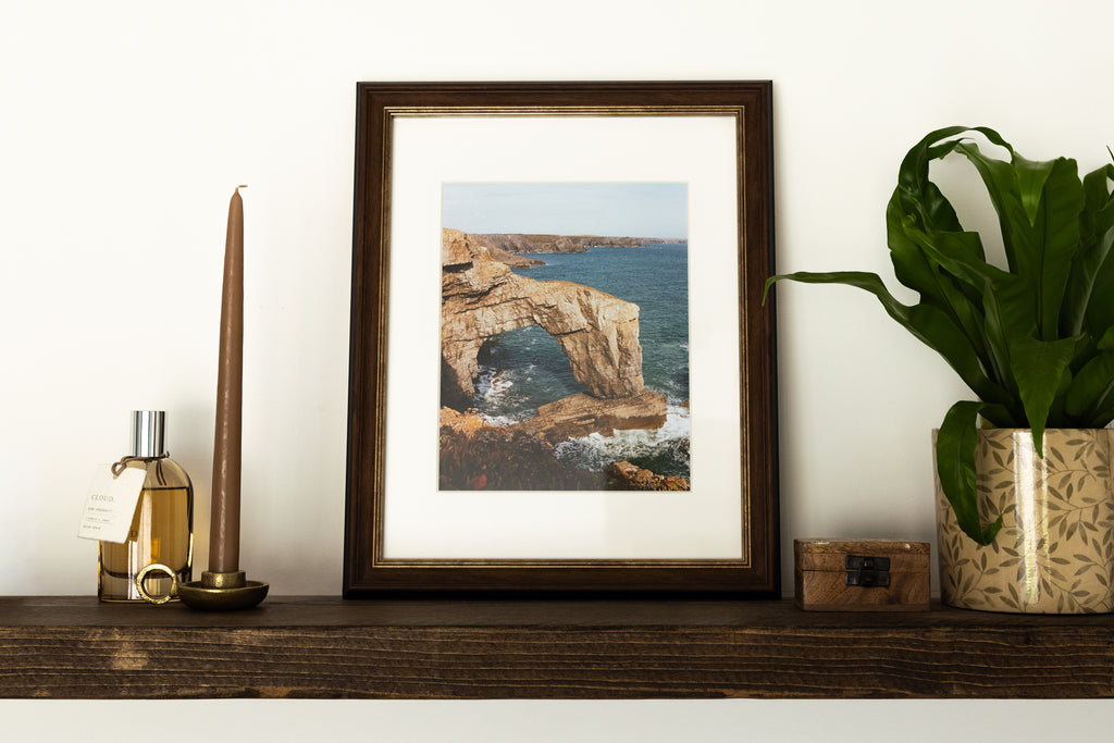 A framed art print depicting a coastal rock arch and sea scene, styled on a dark wooden shelf next to a candle, a small bottle, and a potted green plant against a white wall.