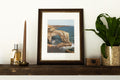 A framed art print depicting a coastal rock arch and sea scene, styled on a dark wooden shelf next to a candle, a small bottle, and a potted green plant against a white wall.