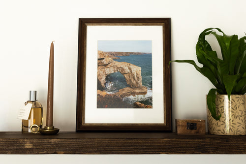 A framed art print depicting a coastal rock arch and sea scene, styled on a dark wooden shelf next to a candle, a small bottle, and a potted green plant against a white wall.