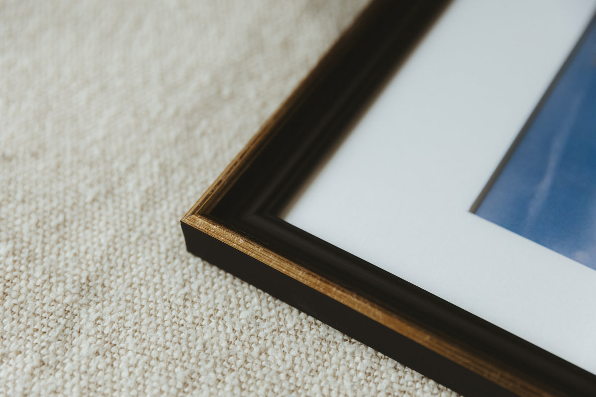 Close-up of a framed art print corner on a textured beige carpet. The frame is dark brown wood with a gold inner trim, set against a white mount and a blue-toned print.