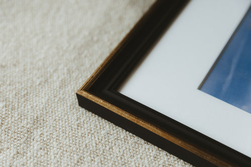Close-up of a framed art print corner on a textured beige carpet. The frame is dark brown wood with a gold inner trim, set against a white mount and a blue-toned print.