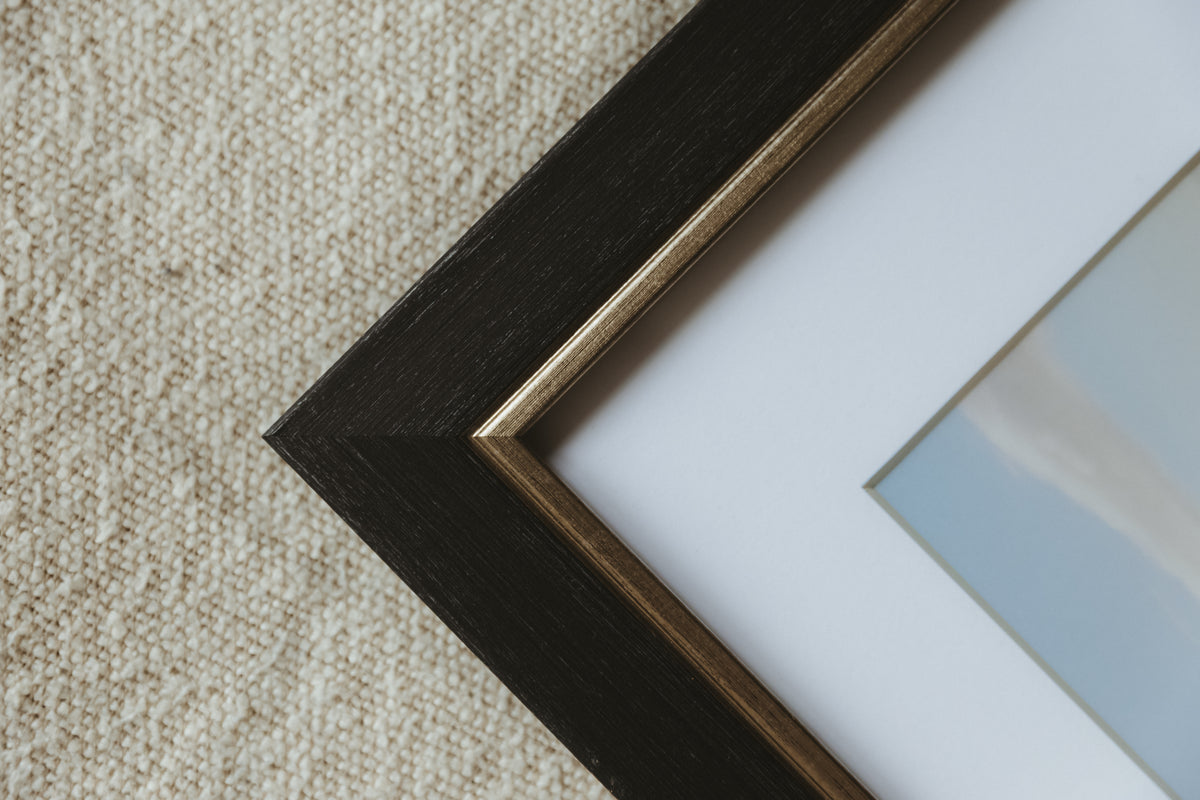 Close-up of a framed art print corner, showing a dark brown wood frame with a gold inner trim, white matting, and a light blue and white print, set against a beige textured rug.