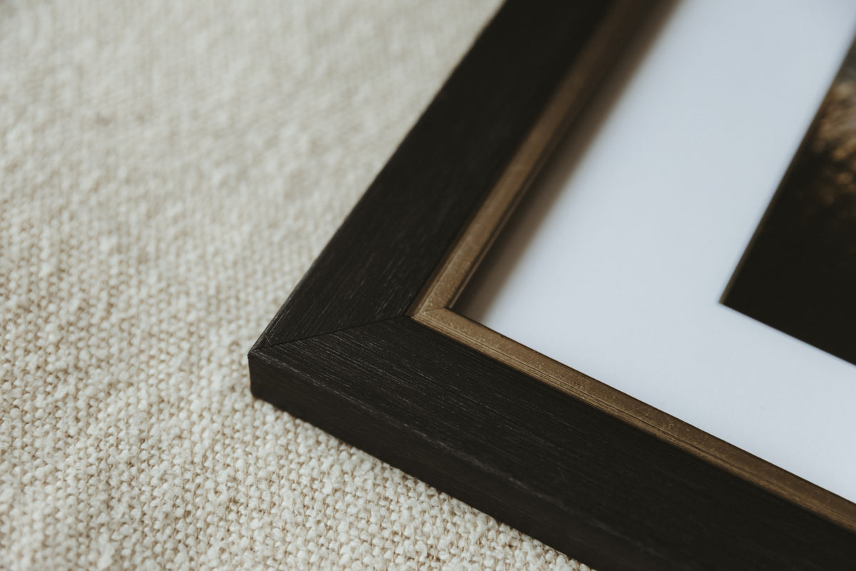 Close-up of a dark wood picture frame corner with a white mount, resting on a textured beige fabric surface.
