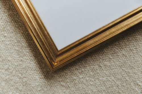Close-up of a decorative picture frame corner with a textured gold finish, resting on a beige woven fabric surface.