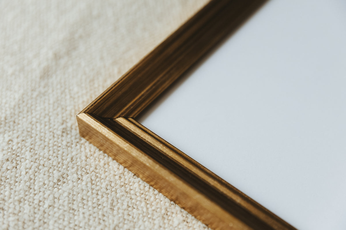 Close-up of a square picture frame corner with a warm medium-brown wood finish, set against a textured beige fabric background.