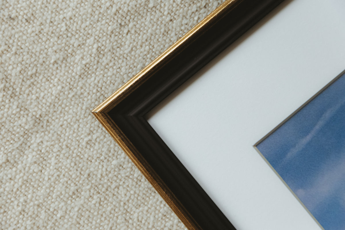 Close-up of a framed art print corner on a textured beige carpet. The frame is black with a gold inner trim, surrounded by a white mount and a blue-toned print.