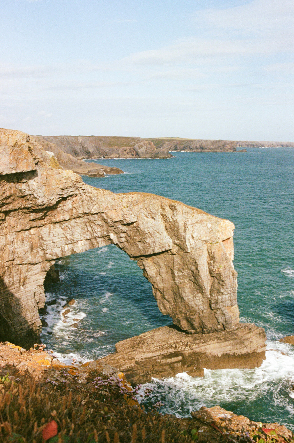A coastal landscape photograph showing a large natural rock arch over blue-green ocean water, with waves crashing at the base and distant cliffs under a pale sky.